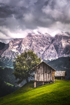 Wooden cottages near a tree on a grassy hill with mountains in the background and dark cloudy sky in Dolomites, Italy