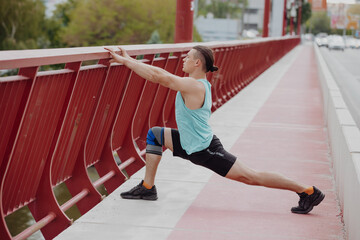 Athlete man stretches leg before running on a bridge in city