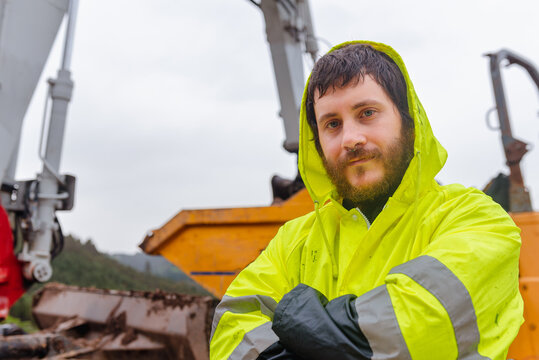 portrait of laborer with piercing, beard and reflective jacket with construction machinery in the background. man with arms crossed and looking at camera.