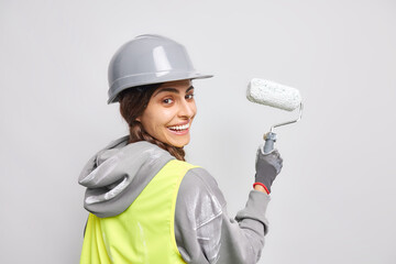 Positive happy female worker in hardhat and uniform paints wall with roller uses instruments or tools for apartment renovation busy doing redecoration poses indoor. House improvement concept