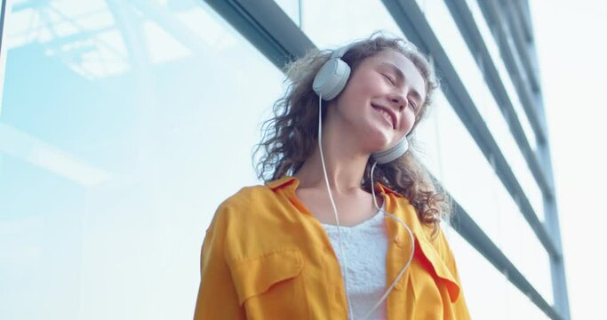 Close up shot of Caucasian cheerful young female dancing outdoor listening to fave song outdoors. Beautiful woman with smile on face enjoys music standing on street in town. Leisure, relax concept