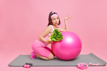 Obraz na płótnie Canvas Fitness at home. Confused hesitant thoughtful Asian sportswoman poses with fitness ball green vegetable motivates you to healthy lifestyle does pilates exercises isolated over pink background