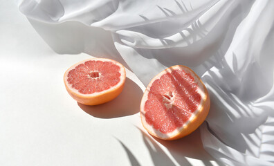 Close up summer still life flat lay with two cut grapefruits and white silk fabric on table in sunlight. Palm leaves harsh shadows. Minimal concept for healthy lifestyle, dieting, freshness. Vacation
