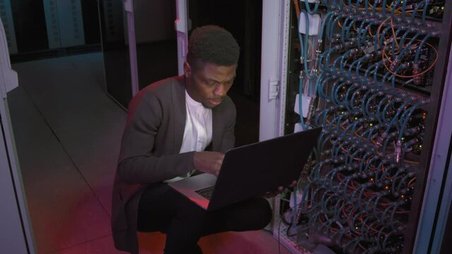 Tracking shot of male African-American network engineer crouching before rack with switches or other computing equipment and typing on laptop