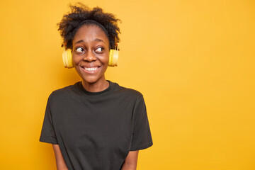 Glad dark skinned woman smiles happily looks away wears headphones on ears dressed in black t shirt isolated over yellow background with copy space area. Music is my life. People and hobby concept