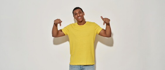 Smiling young man in casual yellow t shirt looking at camera and pointing with index fingers down while presenting your product, standing isolated over gray background