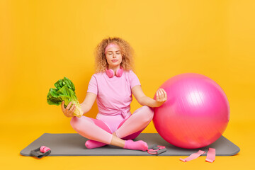 Fototapeta na wymiar Athletic young woman sits in lotus pose tries to relax meditates after fitness training practices yoga holds green vegetable sits on mat near fitball sport equipment isolated over yellow background