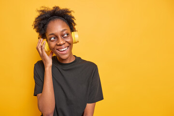 Studio shot of happy millennial girl with curly hair keeps hand on headphones listens music smiles happily looks away dressed in black casual t shirt isolated over yellow background copy space
