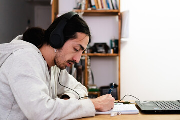 young latino man studying at home, with a notebook and a computer, listening to music with headphones.