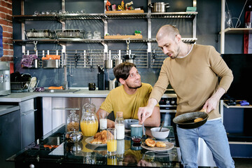 Happy young man looking at his boyfriend with frying pan during breakfast