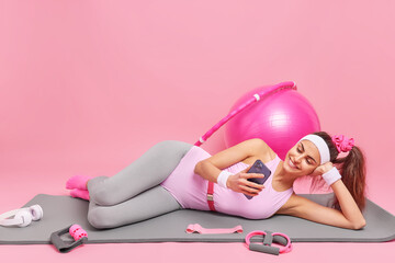 Obraz na płótnie Canvas Horizontal shot of relaxed young brunette woman lies on comfortable fitness mat watches video via smartphone dressed in sportswear exercises with sport equipment isolated over pink background