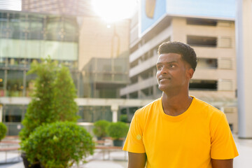 Fototapeta premium Portrait of handsome black African man wearing yellow t-shirt outdoors in city during summer backlit shot with lens flare while thinking