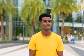 Portrait of handsome black African man wearing yellow t-shirt outdoors in city during summer while thinking