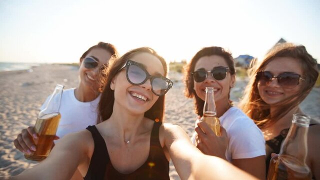 Pov view of pretty women take selfie having fun with drinks on sea beach on sunset. Online video call: girl looking at smartphone camera on tropical island, females toasting bottles, waving hands.