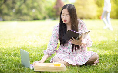 Working outside in summer or spring and WFH concept, Young beautiful Asian woman reading a book and looking laptop in the natural park, Freelance working, homeschooling, distance learning.