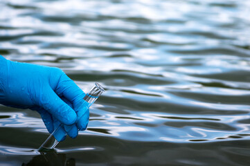 A gloved hand holds a test tube of clear water against the background of the waves of the reservoir. Background.
