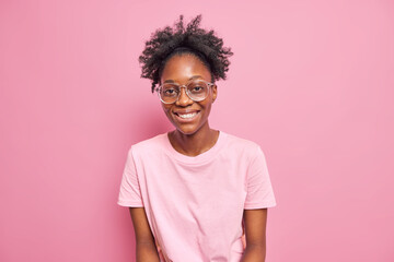 Fototapeta na wymiar Skinny dark skinned woman with curly hair smiles broadly enjoys leisure free time wears big transparent glasses t shirt isolated over pink background. Peope ethnicity sincere emotions concept