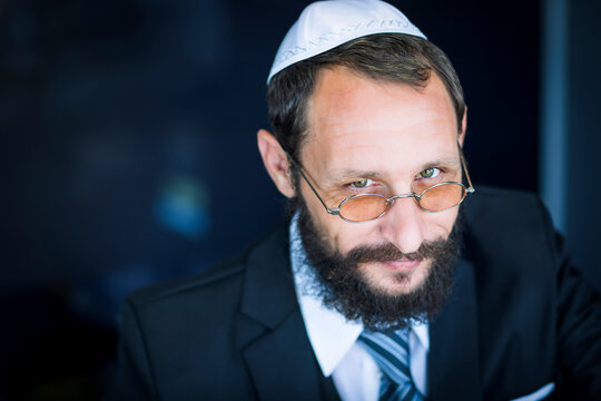 Bearded Charming Jewish man in white Yarmulke (hat, Kippah) looking with cunning eyes. Emotional expression. Sly bearded Jewish man smiling cunning looking at the camera. Selective focus on the eyes