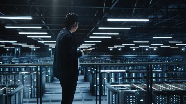 Portrait of IT Specialist Using Tablet Computer in Data Center. Big Server Farm Cloud Computing Facility with Male System Administrator Working. Cyber Security, Network Protection. Full Wide Arc