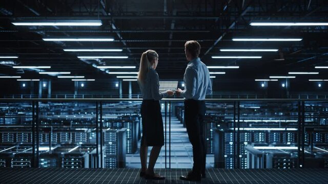 Data Center Male It Specialist and Female Maintenance administrator Talk, Use Laptop. Server Farm Cloud Computing Facility with Two Information Technology Professionals working on Cyber Secury. Wide