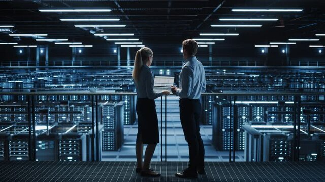 Data Center Male It Specialist and Female System administrator Talk, Use Laptop. Server Farm Cloud Computing Facility with Two Information Technology Professionals working on Cyber Secury. Static Wide