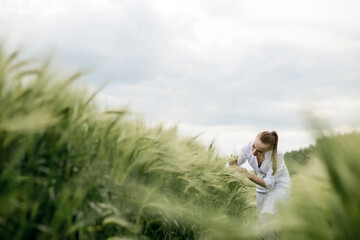 Naklejka na ściany i meble Young woman agronomist in white coat squatting in green wheat field and checking crop quality