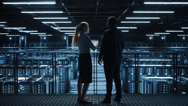 Data Center Male It Specialist and Female System administrator Talk, Use Laptop. Big Server Farm Cloud Computing Facility with Information Technology Professionals working. Pedestal Wide Shot