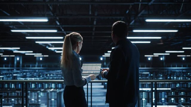 Data Center Male It Specialist and Female e-Commerce Manager Talk, Use Laptop. Big Server Farm Cloud Computing Facility with Two Information Technology Professionals working. Medium Wide Shot