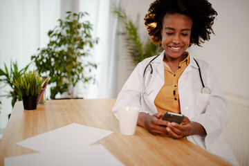 Female Afro American doctor sitting with mobile phone and drinking coffee.