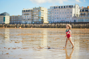 Adorable toddler girl in red swimsuit playing with wooden toy boat in water on the beach in Saint-Malo, Brittany, France