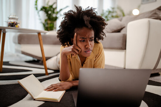 Portrait Of Worried Young African American Woman Using Laptop While Lying On Floor At Home.