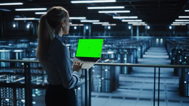 Female It Specialist Using Chroma Key Laptop in Data Center. Big Server Farm Cloud Computing Facility with Maintenance Engineer Working with Green Screen Computer. Medium Wide over the shoulder