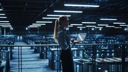 Data Center Female IT Specialist Uses Laptop Computer. Information Technology Engineer Overseeing and Walking Through Cloud Computing Server Farm Maintenance and Monitoring. Profile Medium Shot
