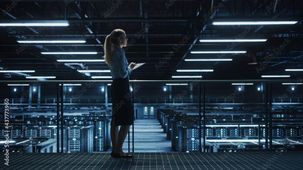 Poster female it specialist using tablet computer in data center, walking on a bridge overlooking big serve