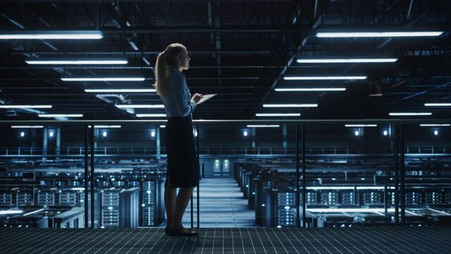 Female IT Specialist Using Tablet Computer in Data Center, Walking on a Bridge Overlooking Big Server Farm Cloud Computing Facility. Businesswoman, e-Business Entrepreneur. Pedestal Wide Shot - Powered by Adobe