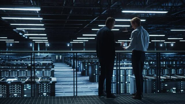 Data Center IT Specialist and e-Business Enrepreneur Talk, Use Laptop. Two Information Technology Professionals Walking on Bridge Overlooking Cloud Computing Server Farm. Medium Wide Shot