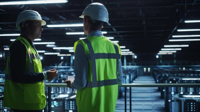 Data Center IT Specialist and System administrator Talk, Use Tablet Computer, Wearing Safety Wests. Server Clod Farm with Two Information Technology Engineers checking Cyber Security. Medium Wide
