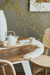 Interior design of stylish dining room interior with family wooden and epoxy table, rattan chairs, flowers in vase and teapot with cups.  Details. Template.