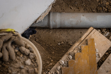 Leaking plastic sewage pipe in an old apartment, visible open hole with removed parquet, sand and buckets around, exposed drain pipe in the ground.