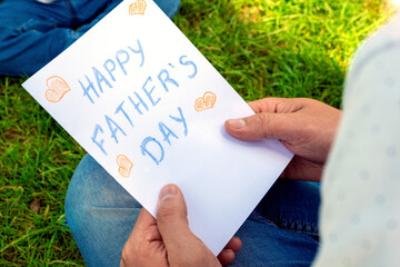 Man sit on grass and hold in hands Happy Father`s Day greeting card as surprise present from kid. Child`s handmade postcard crafts. Family, togetherness, love, relationships. Happy Father`s Day.