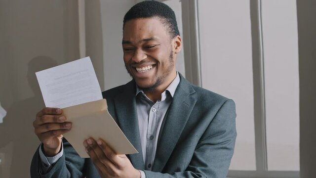 Smiling African stylish young guy black man businessman in modern office room alone read letter grand proposal exam results financial report paperwork feels happy celebrate victory. Good news concept