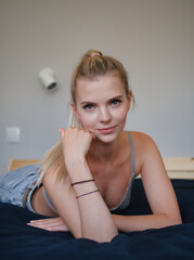 Wonderful day happy smiling blonde young blue-eyed woman having fun on the sofa staring into camera holding her head dressed blue denim jeans grey bra