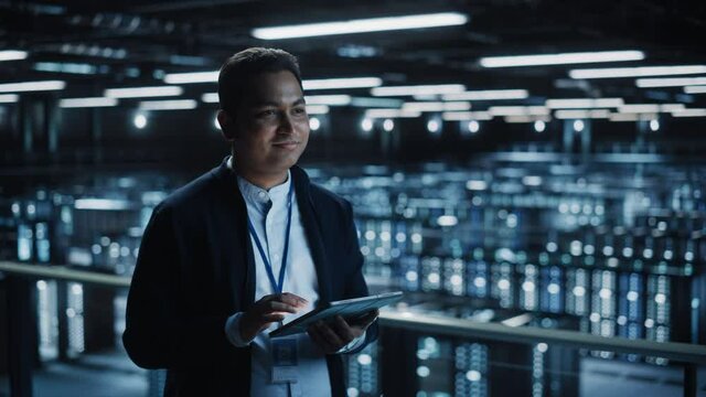 Handsome Smiling IT Specialist Using Tablet Computer in Data Center. Succesful Businessman and e-Business Entrepreneur Overlooking Server Farm Cloud Computing Facility. Medium Wide Shot