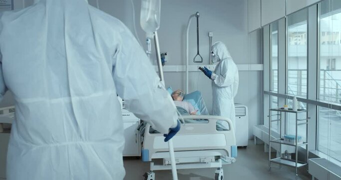Doctor and nurse in ppe overall treating aged woman patient with covid-19 in hospital ward