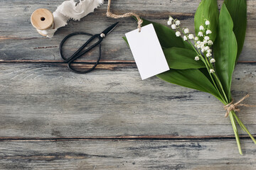 Spring decorative banner. Blank gift tag label mockup, black vintage scissors. Silk ribbon and bouquet of blooming lilly of the valley flowers on old wooden table background. Flat lay, top view.