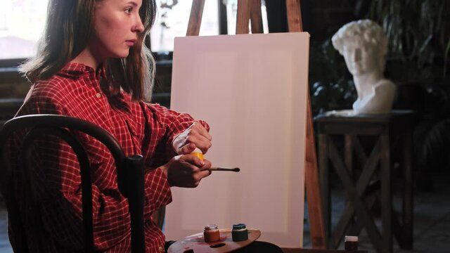 A young woman in art workshop opens up jars of color paints sitting on front of an easel