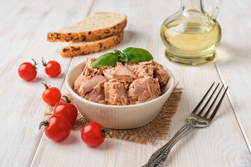 Canned tuna meat in a bowl, fork, bread and fresh red cherry tomatoes on a white wooden table. Low calories healthy eating snack of preserved tuna fish and vegetables. Tasty seafood.
