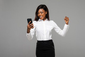 Excited African emotional woman talking on phone happy to hear good news clenched fist achieved desired posing isolated on grey background