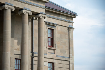 Fototapeta premium A wide brick entrance to a large courthouse, justice or government style building. There's a tall red door, grey marble steps, black handrails, tall windows, round pillars and a beige exterior wall. 