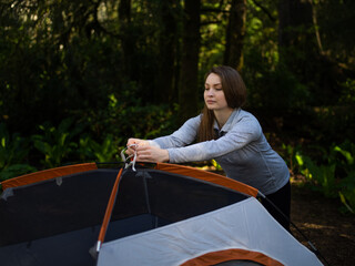 In the photo, a young woman, a tourist lays out a tent in a tent camp. Around a dense green forest. Evening time. Dusk. Muted tones. Lots of greenery. The tent is blue and brown.